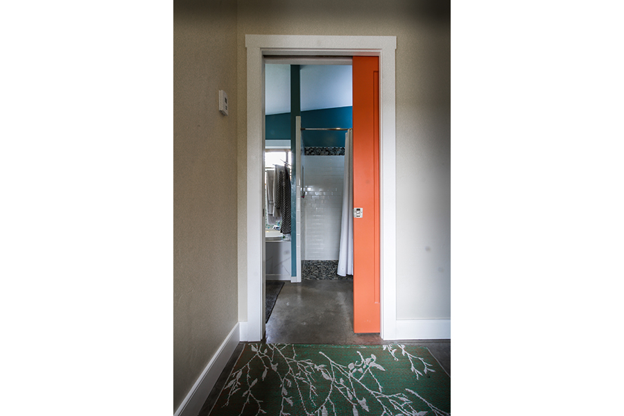 A green rug gives way to an orange pocket door, sliding into a wall. The entrance leads to a bathroom, which has a shower that is seemlessly accessible from bathroom floor with no threshold.